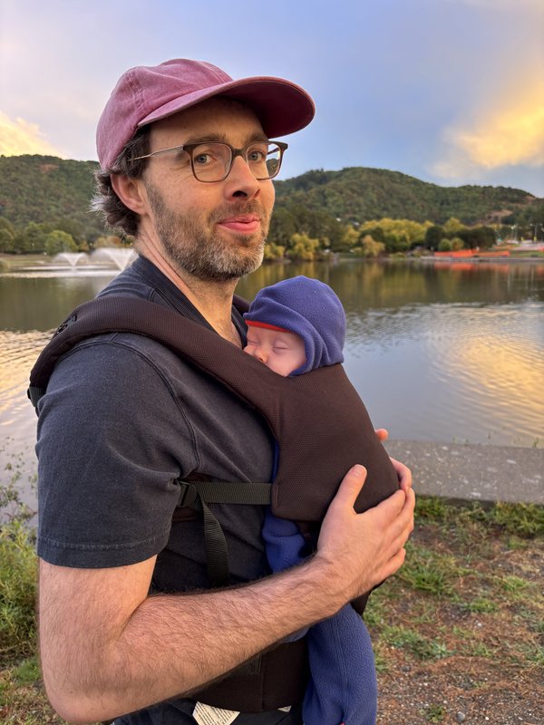 Paul with his son Wallace in a baby carrier, beside a lake at sunset